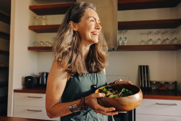 A woman in a kitchen holds a wooden bowl of salad, smiling happily, conveying a feeling of contentment to healthy eating.
