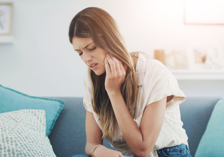 A young woman sitting on a couch holding her cheek and jaw, clearly indicating tooth pain.