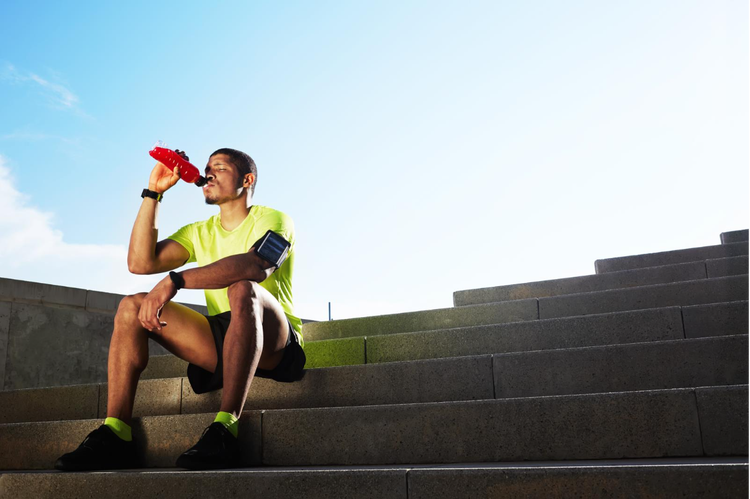 An athlete in a neon green shirt and black shorts sits on concrete stairs outdoors on a sunny day, drinking from a water bottle to stay hydrated in the heat.