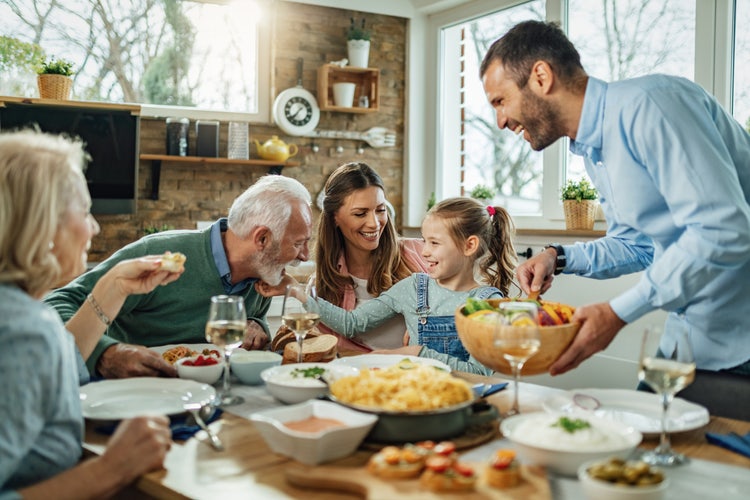 A smiling family of multiple generations is gathered around a dining table. A man on the right is serving a bowl of fresh vegetables.
