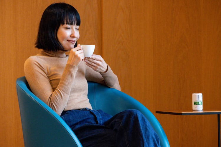 A woman sitting in a room, smiling as she drinks a healthy Sunstar product.